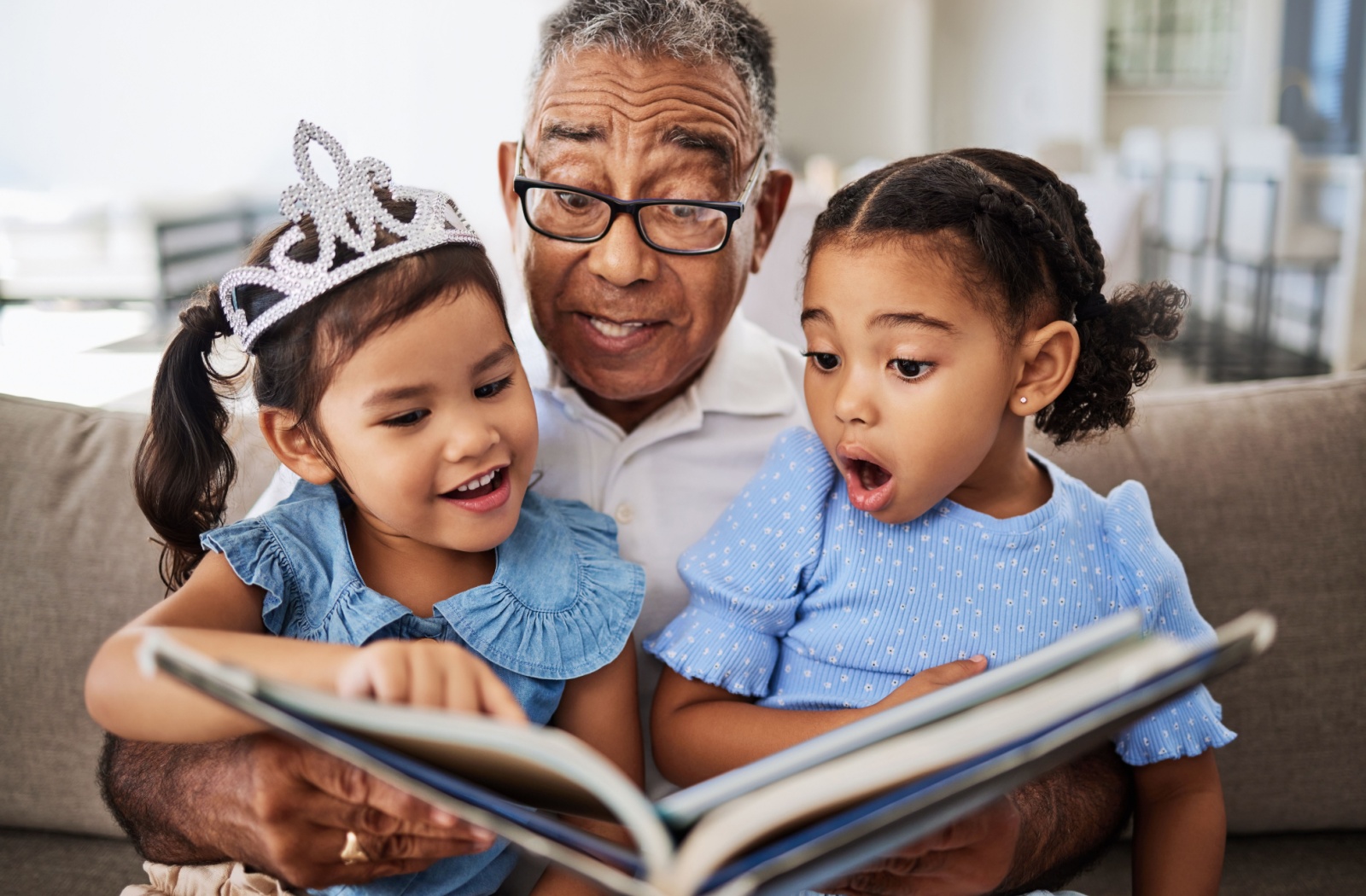 A senior man reads a book with his granddaughters. Each of them wears a delighted expression.