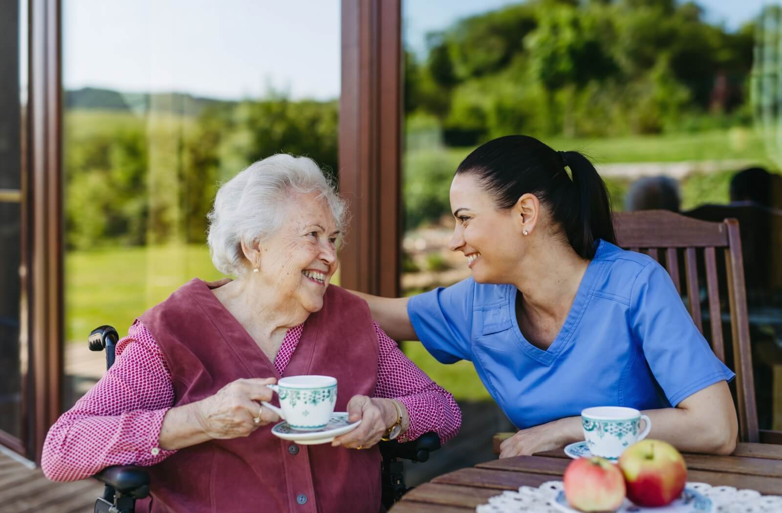 A caregiver and a resident in assisted living sitting in a sunroom and enjoying cups of tea.