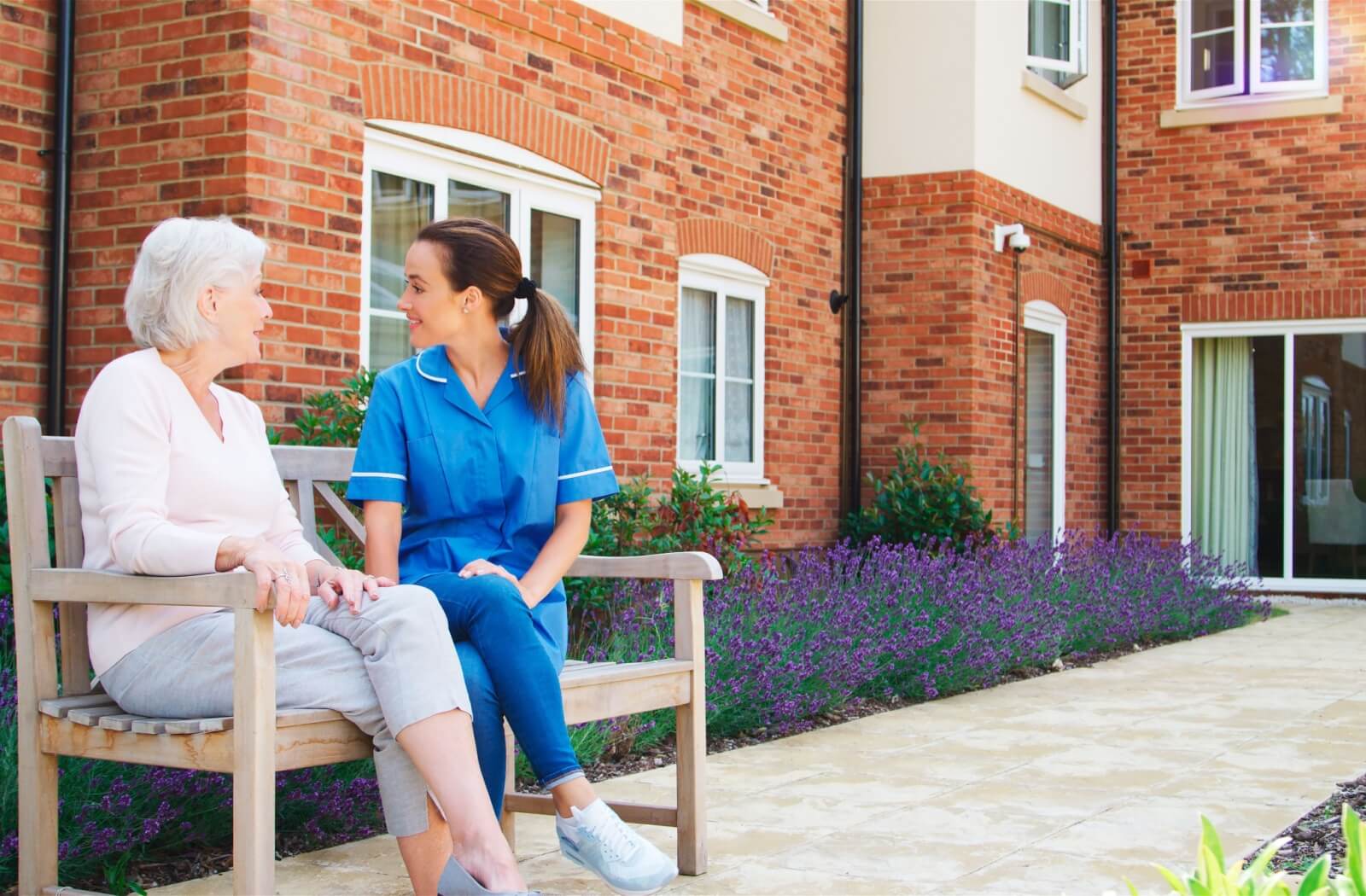 A senior and a nurse sit on a bench, chatting while enjoying the fresh air and beautiful outdoor spaces in assisted living